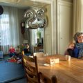 Mariet v.d. Toorn in woonkamer aan eettafel met kind op schoot en erachter spelende kinderen op grond en met 2 zilverkleurige balonnen, 1 met cijfer 2 en een met 5