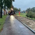 Fietspad rechts van rij bomen, rechts van fietspad ligt de stoep open met zand erin. Werkmannen met bulldozer zijn aan het werk.