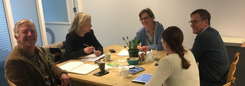 5 personen zitten aan een tafel en hebben overleg