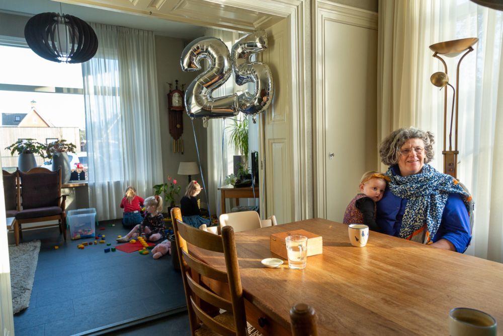Mariet vd Toorn aan haar keukentafel. Op schoot een kind en in de achtergrond zijn ook kinderen aan het spelen met lego. In beeld staan zilveren ballonnen die het getal 25 vormen.
