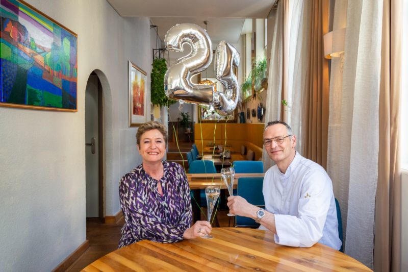 Nancy en Roberto Hoeppe zitten in het restaurant De Eetgelegenheid en heffen het glas. Op de achtergrond zilveren ballonnen in de vorm van het getal 25.